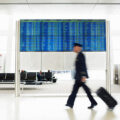 A blurred pilot walks past a board showing departures as he rolls a suitcase.