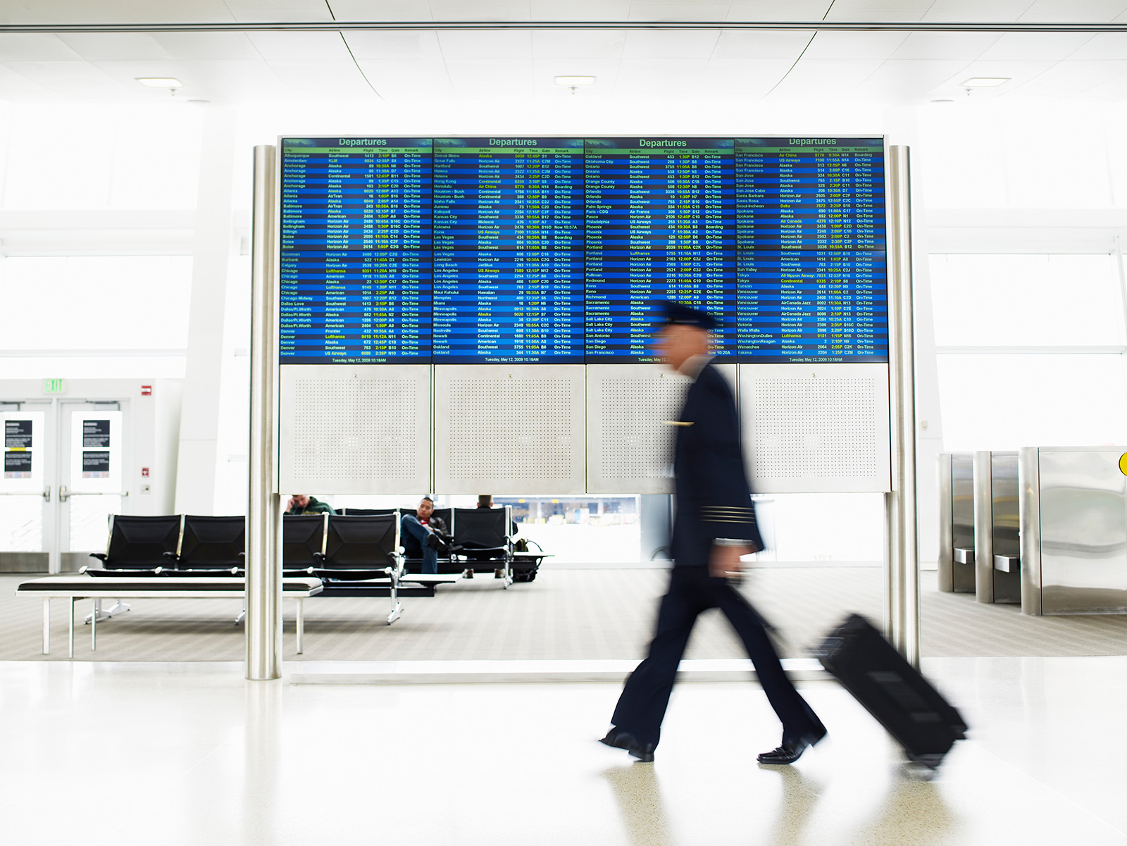 A blurred pilot walks past a board showing departures as he rolls a suitcase.