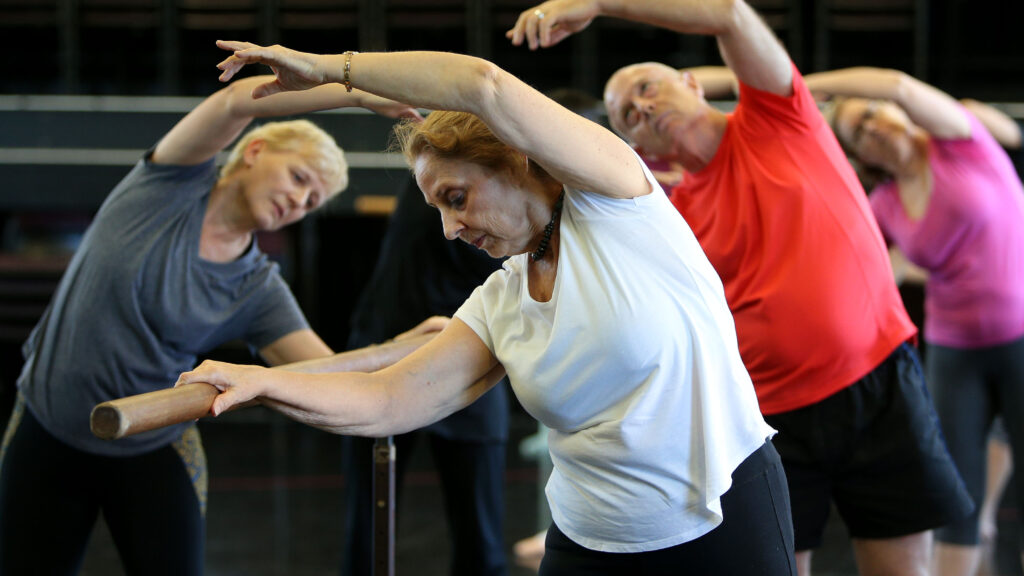 Older women and men practice ballet at a bar.