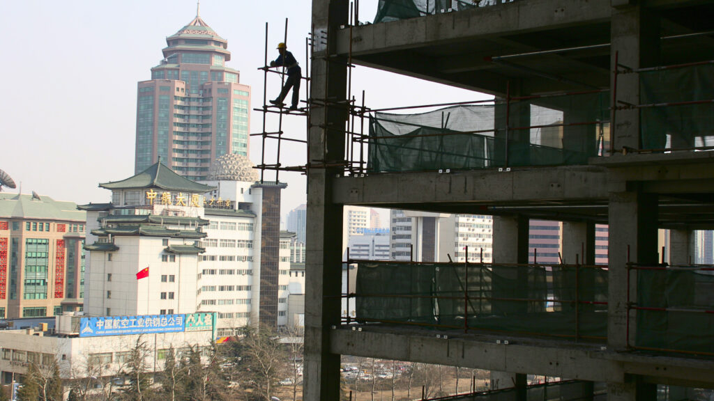 A man stands on scaffolding next to a construction site. In the background are tall buildings.