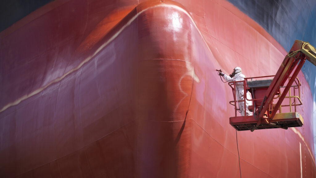 A man in a white protective suit and mask stands on a crane while spraying the hull of a ship red.
