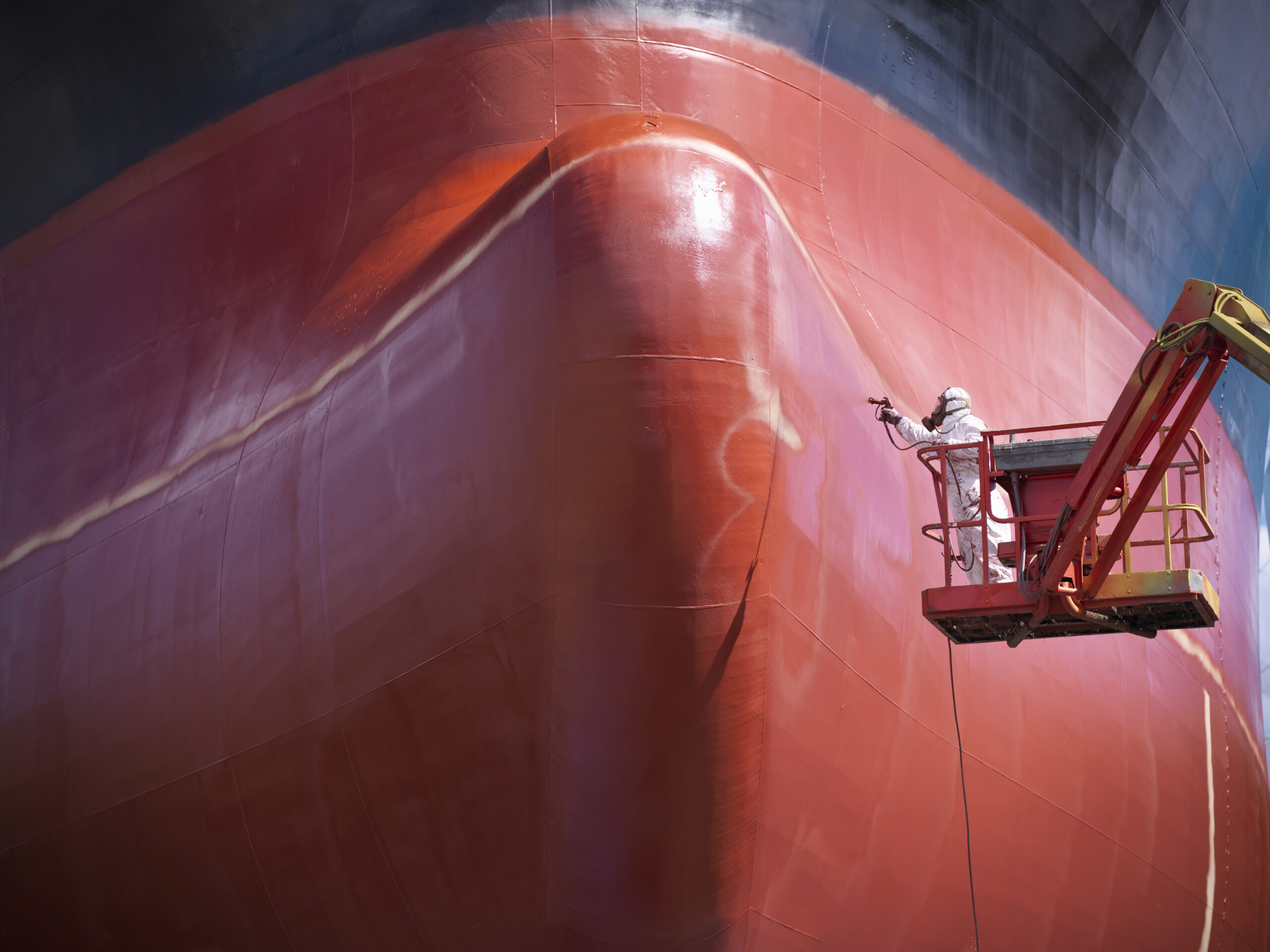 A man in a white protective suit and mask stands on a crane while spraying the hull of a ship red.