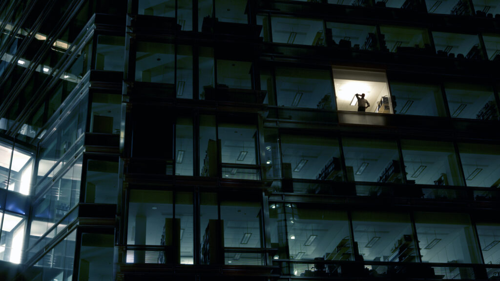 An employee, working late, is seen in an illuminated window of an office building that is otherwise dark.