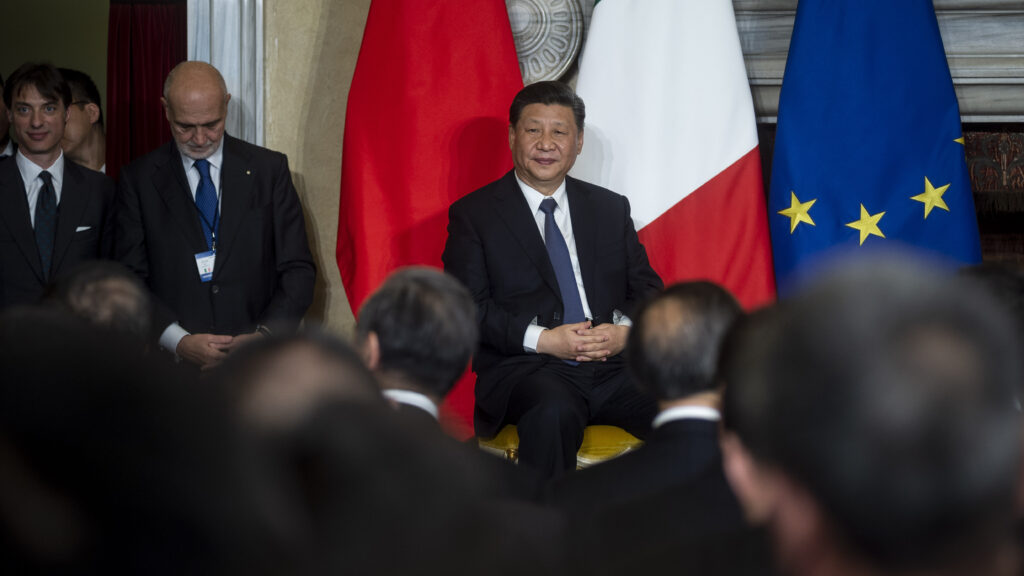 Chinese President Xi Jinping sits in a chair on a stage, wearing a suit and tie. The Chinese, Italian, and EU flags are behind him. A man looking down into his lap stands to Xi Jinping's right. The backs of the heads of a seated crowded are in the foreground.
