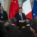 Chinese President Xi Jinping sits in a chair on a stage, wearing a suit and tie. The Chinese, Italian, and EU flags are behind him. A man looking down into his lap stands to Xi Jinping's right. The backs of the heads of a seated crowded are in the foreground.