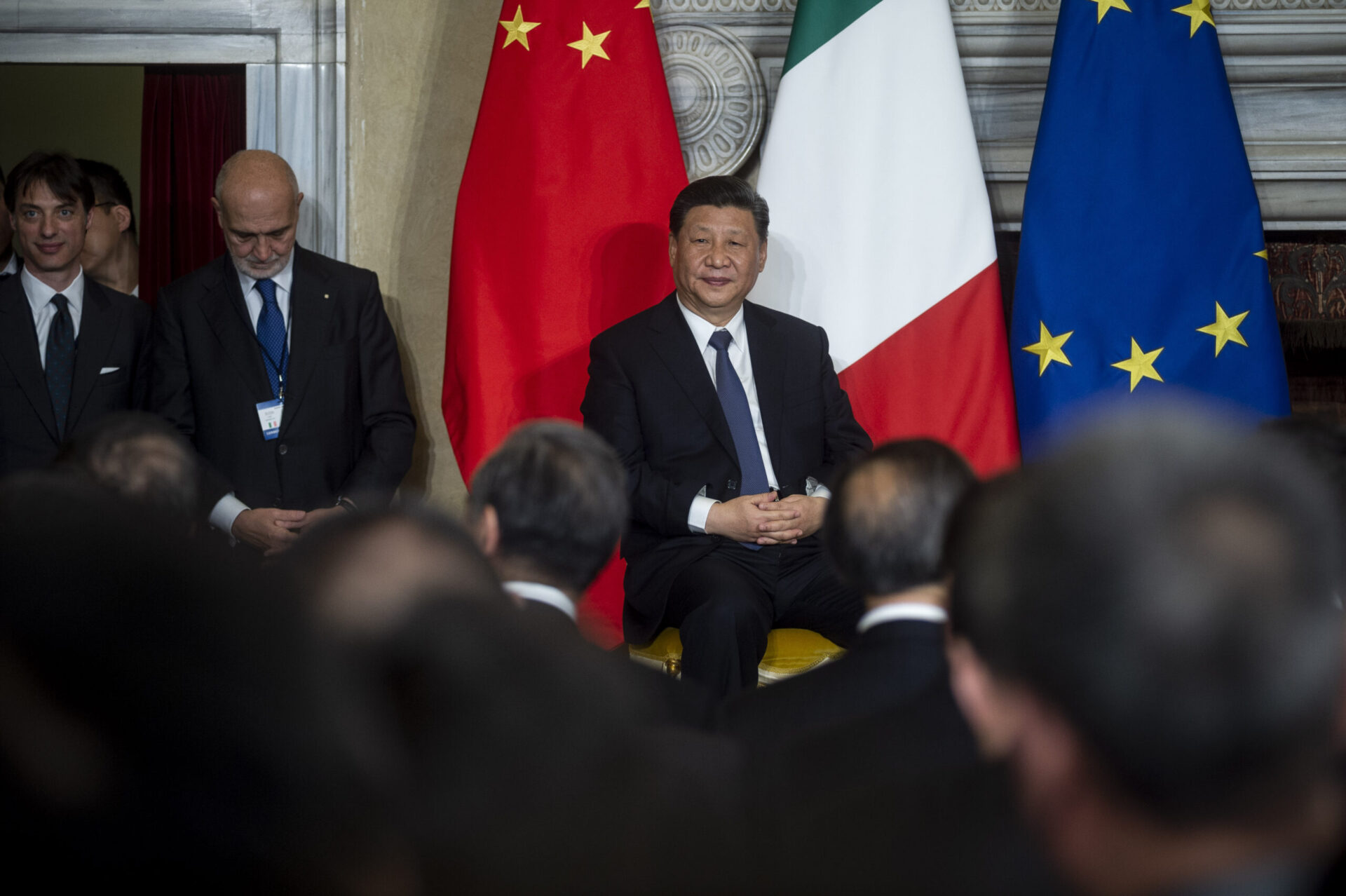 Chinese President Xi Jinping sits in a chair on a stage, wearing a suit and tie. The Chinese, Italian, and EU flags are behind him. A man looking down into his lap stands to Xi Jinping's right. The backs of the heads of a seated crowded are in the foreground.