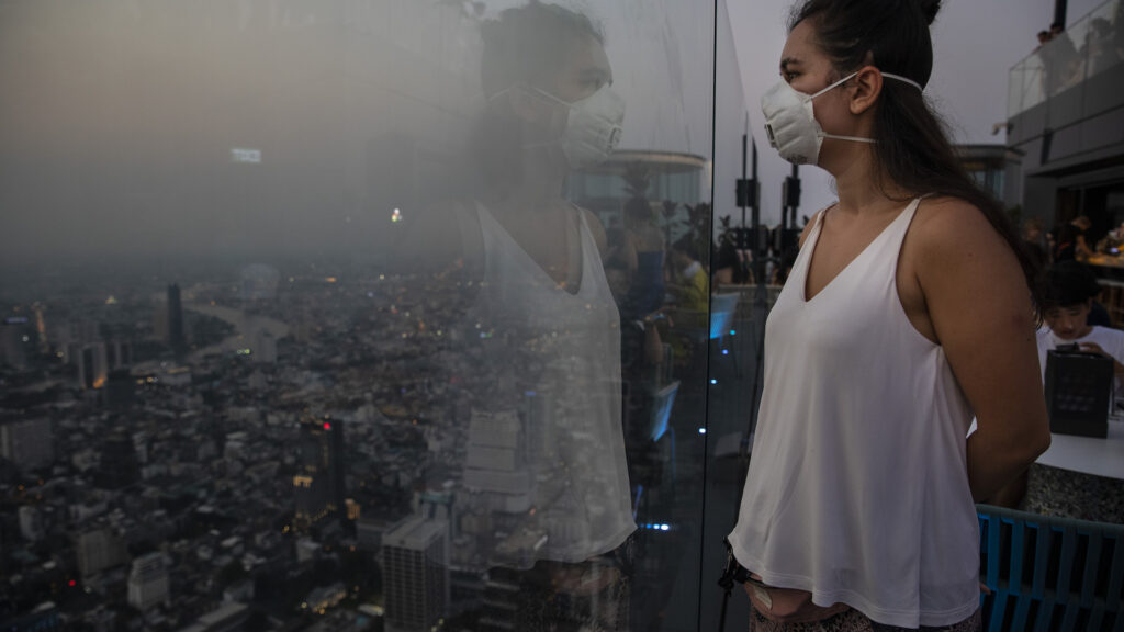 A woman in a white mask and white tank top stands facing a clear glass wall high above a city. The city is shrouded in smog.