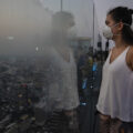 A woman in a white mask and white tank top stands facing a clear glass wall high above a city. The city is shrouded in smog.