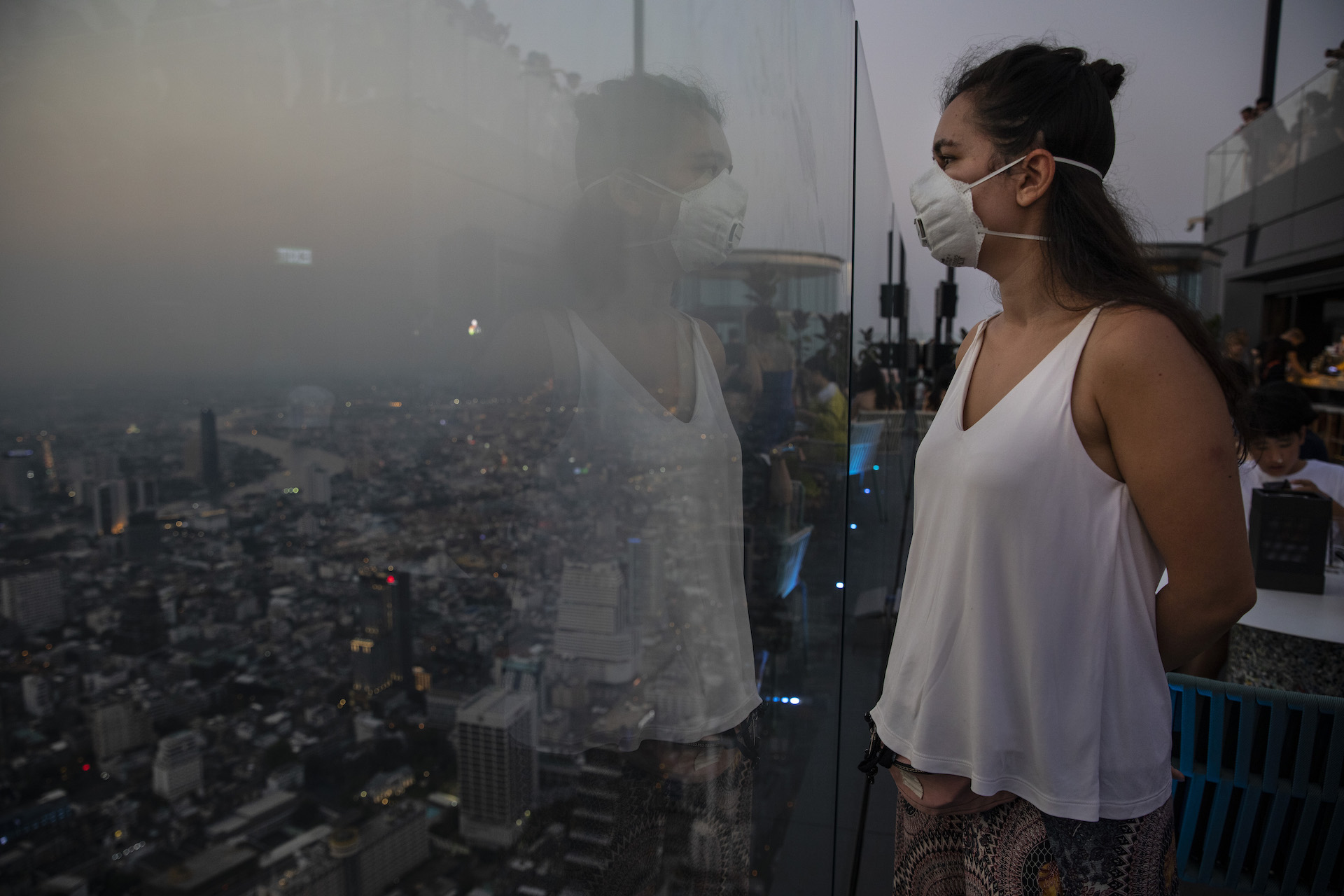 A woman in a white mask and white tank top stands facing a clear glass wall high above a city. The city is shrouded in smog.