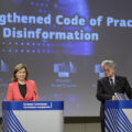 A woman and a man in formal suits stand at two podiums. In the background is a digital wall with the words 