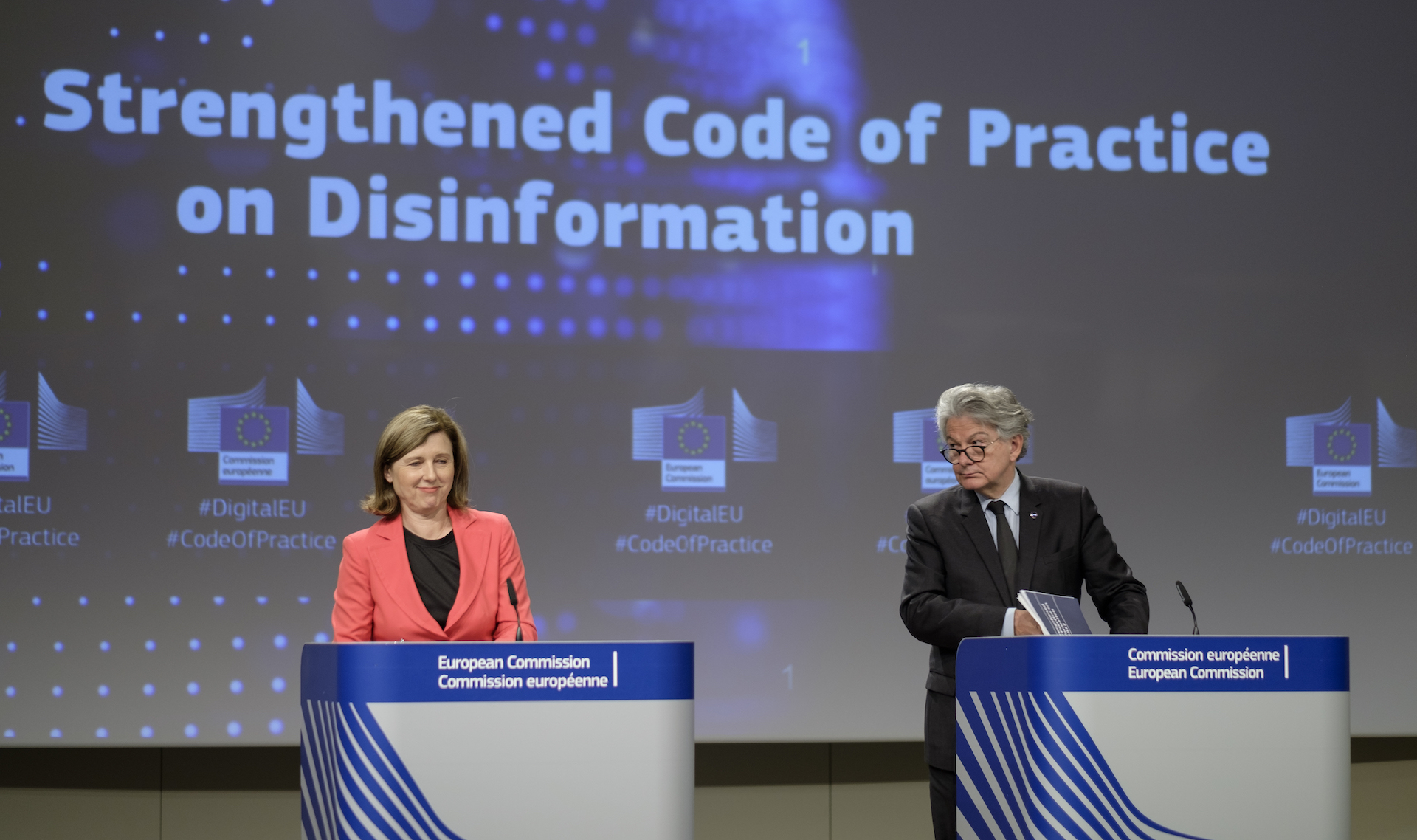 A woman and a man in formal suits stand at two podiums. In the background is a digital wall with the words