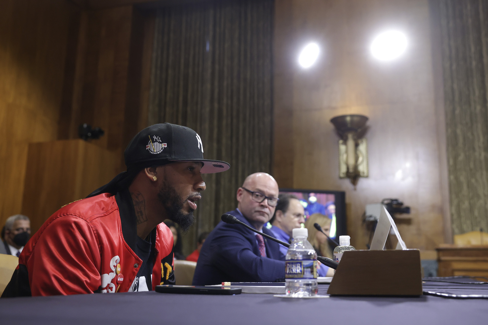 A man in a black baseball hat and orange jacket leans toward a microphone to speak, while sitting at a table. A bald man in a suit and glasses to his left looks at him. They sit in a room with high ceilings and wood paneling.