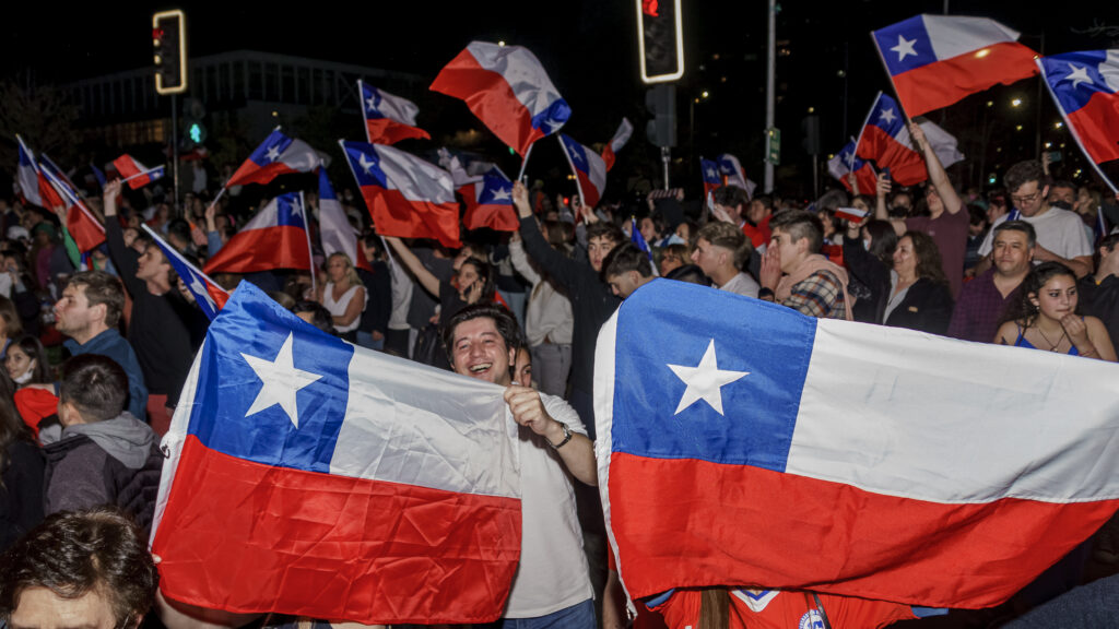 A crowd waves Chilean flags in the street at night. Two giant Chilean flags held up by people take up the foreground, while more flags wave in the background.