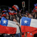 A crowd waves Chilean flags in the street at night. Two giant Chilean flags held up by people take up the foreground, while more flags wave in the background.