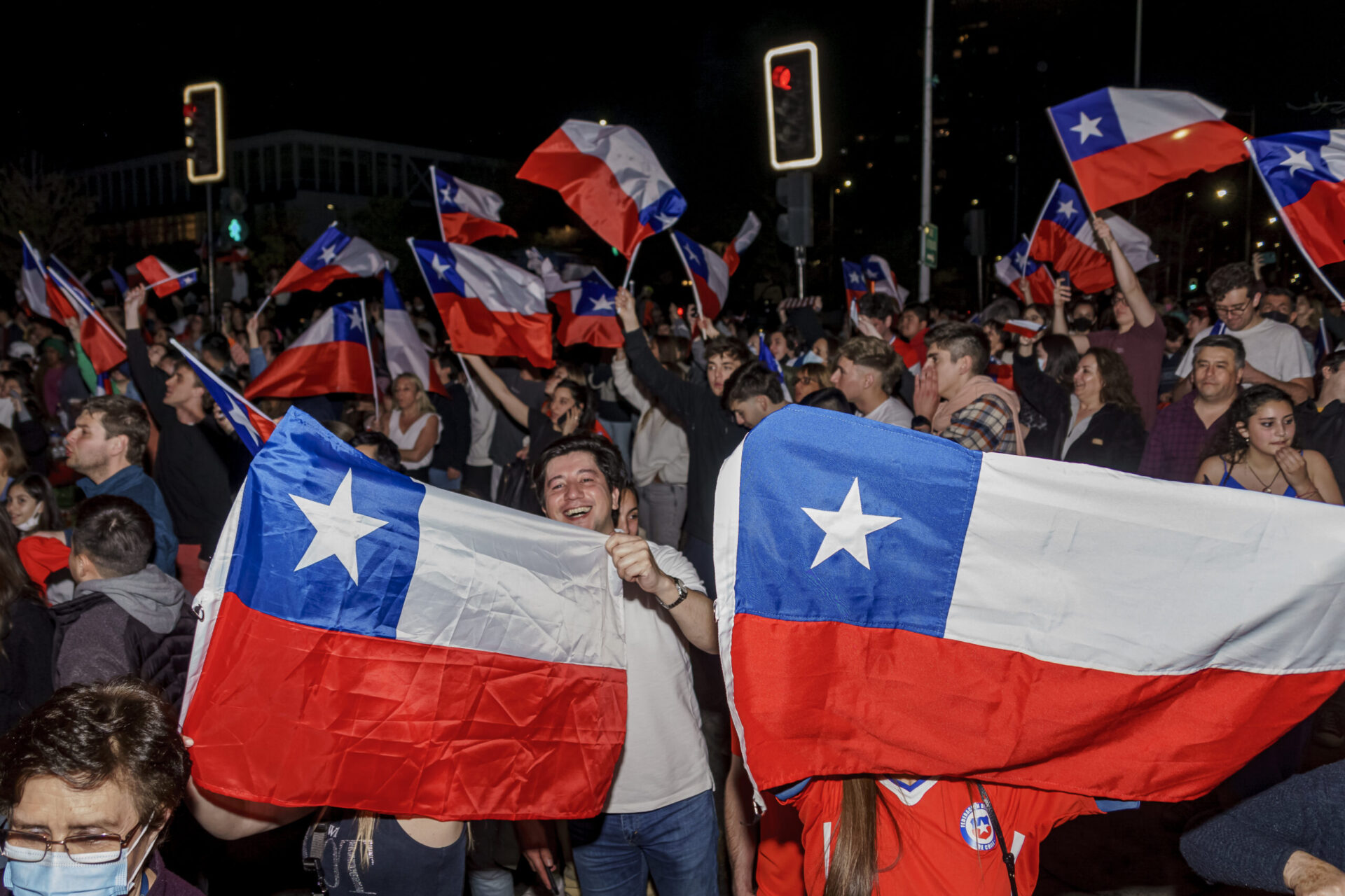 A crowd waves Chilean flags in the street at night. Two giant Chilean flags held up by people take up the foreground, while more flags wave in the background.