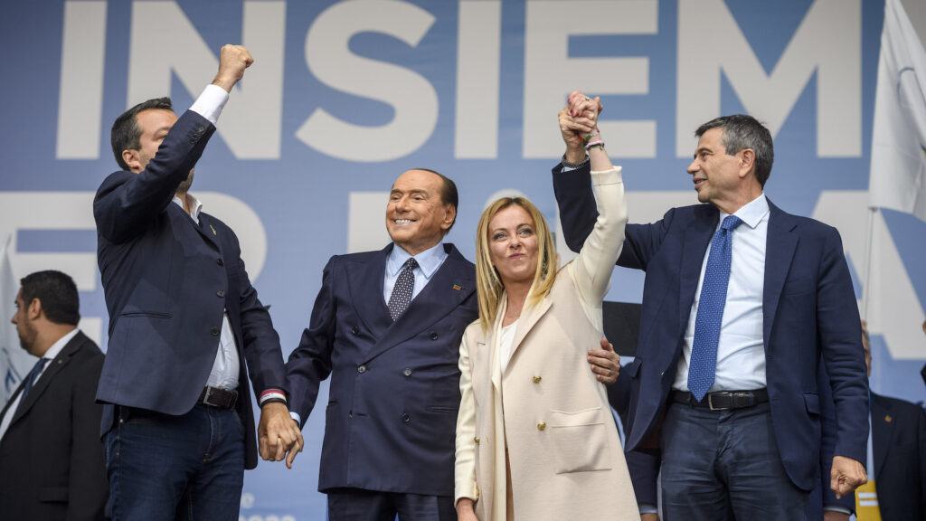 Three men in suits and a woman in an off-white coat hold hands over their head triumphantly. Behind them is a blue background with the word 