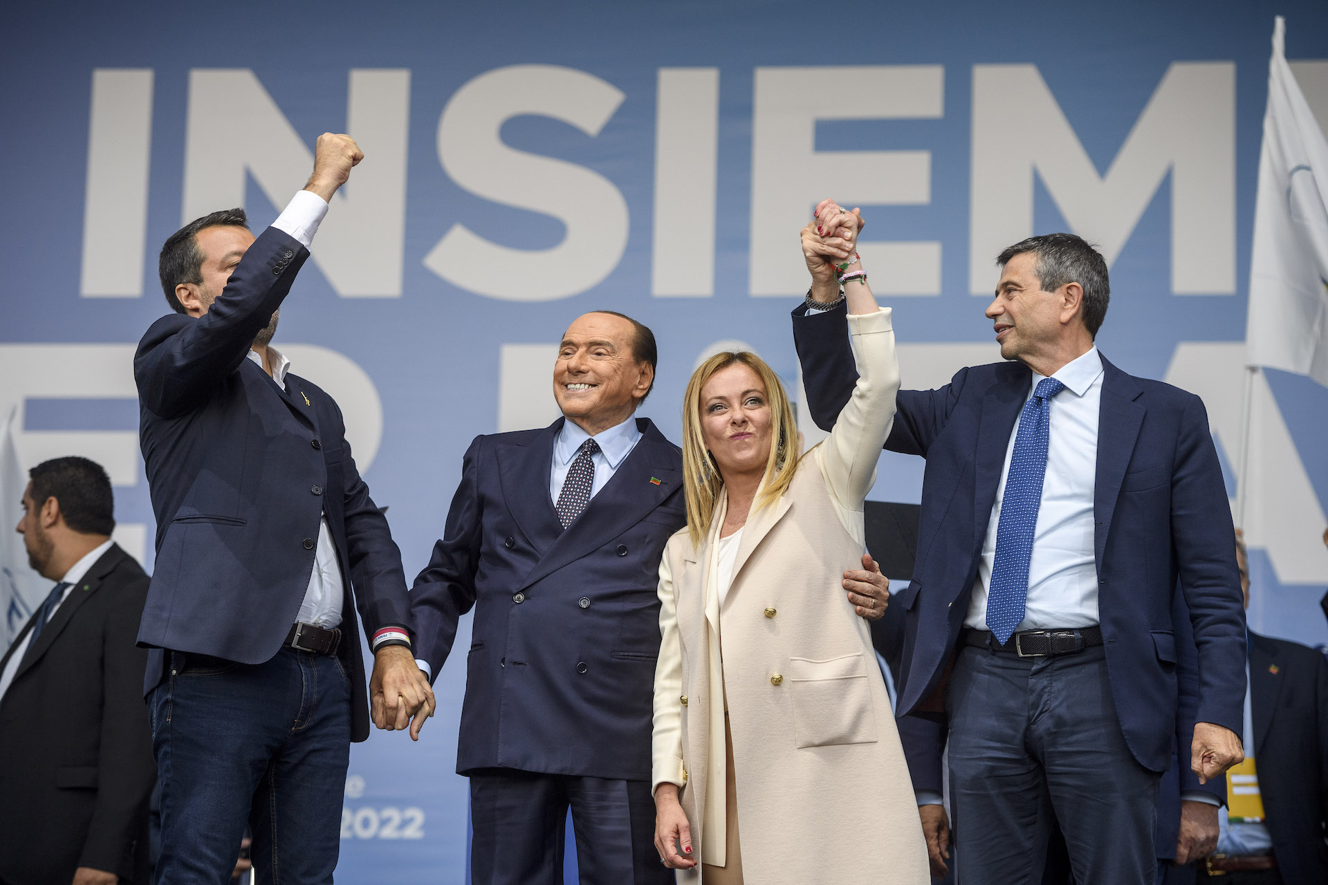 Three men in suits and a woman in an off-white coat hold hands over their head triumphantly. Behind them is a blue background with the word