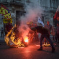 A small crowd in a street at dusk burns large sheets of paper. In the foreground, a man bends at the waste to light a paper on fire; the fire casts a red and orange glow over the picture. Spectators form a semi-circle around the fire, holding union flags that say USB. A building made of stone with carved pillars and balconies is in the background.