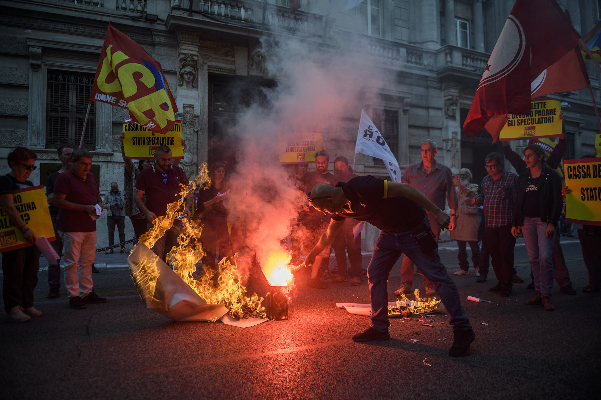 A small crowd in a street at dusk burns large sheets of paper. In the foreground, a man bends at the waste to light a paper on fire; the fire casts a red and orange glow over the picture. Spectators form a semi-circle around the fire, holding union flags that say USB. A building made of stone with carved pillars and balconies is in the background.
