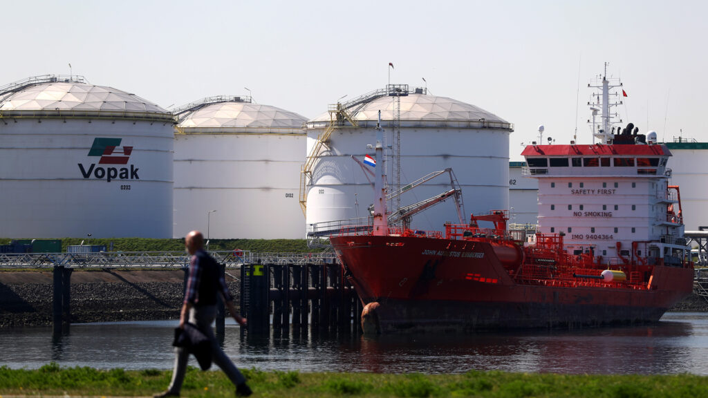 White circular buildings housing oil and gas sit on the water. A man walks by them on the grass on the other side of the bank.