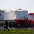 White circular buildings housing oil and gas sit on the water. A man walks by them on the grass on the other side of the bank.