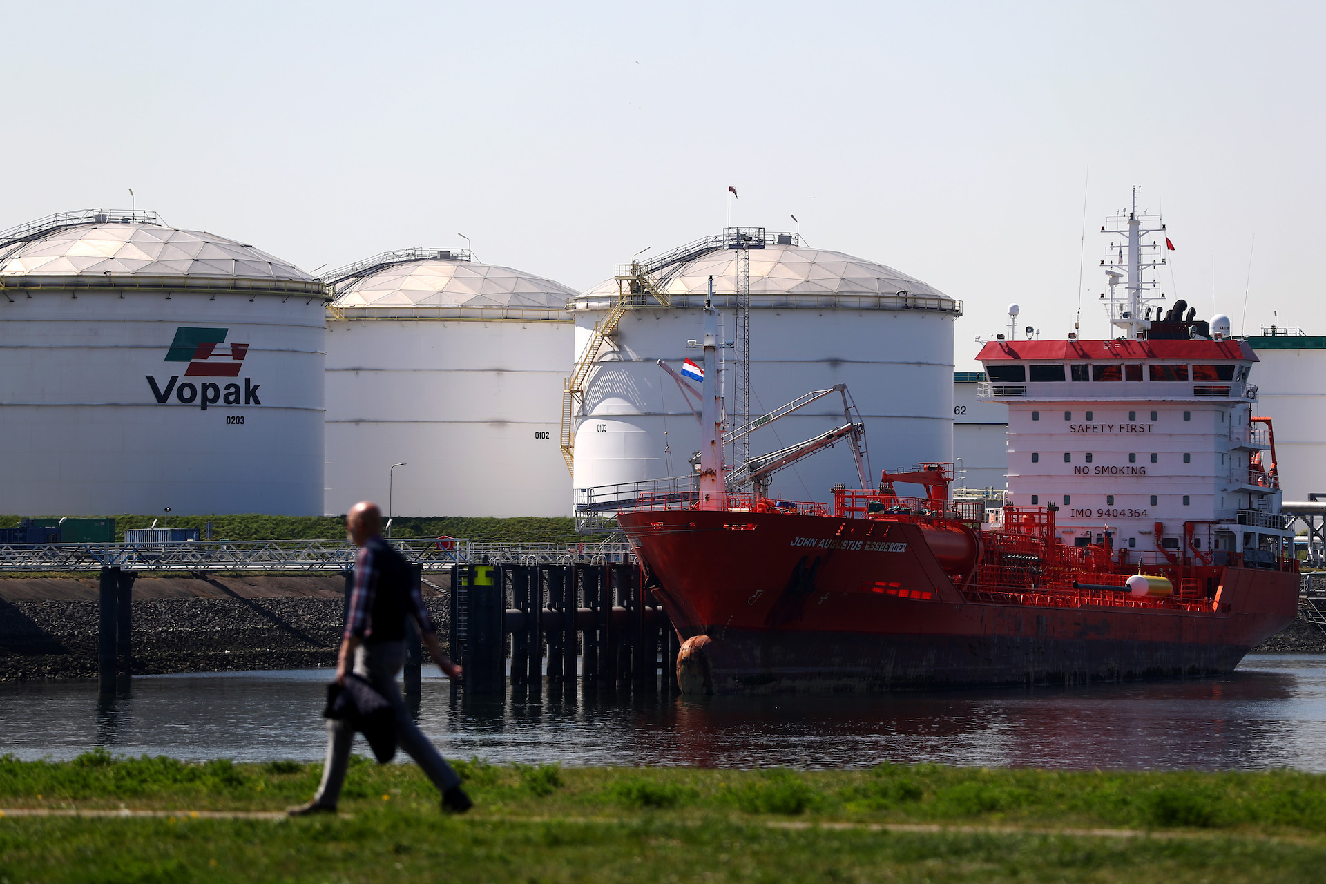 White circular buildings housing oil and gas sit on the water. A man walks by them on the grass on the other side of the bank.