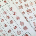 Two white sheets of paper are covered in vertical rows of red circular stamps, with Japanese characters.