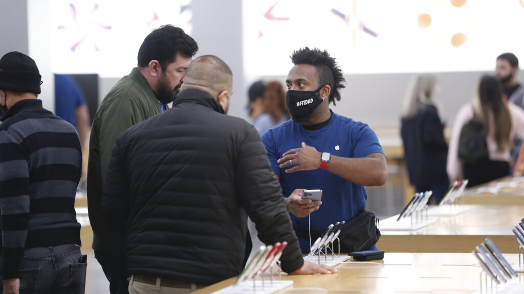 An employee in a blue shirt and black mask gestures as he talks to two male customers in winter jackets. They are at a table holding cell phones, with a white lit wall behind them.