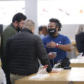 An employee in a blue shirt and black mask gestures as he talks to two male customers in winter jackets. They are at a table holding cell phones, with a white lit wall behind them.