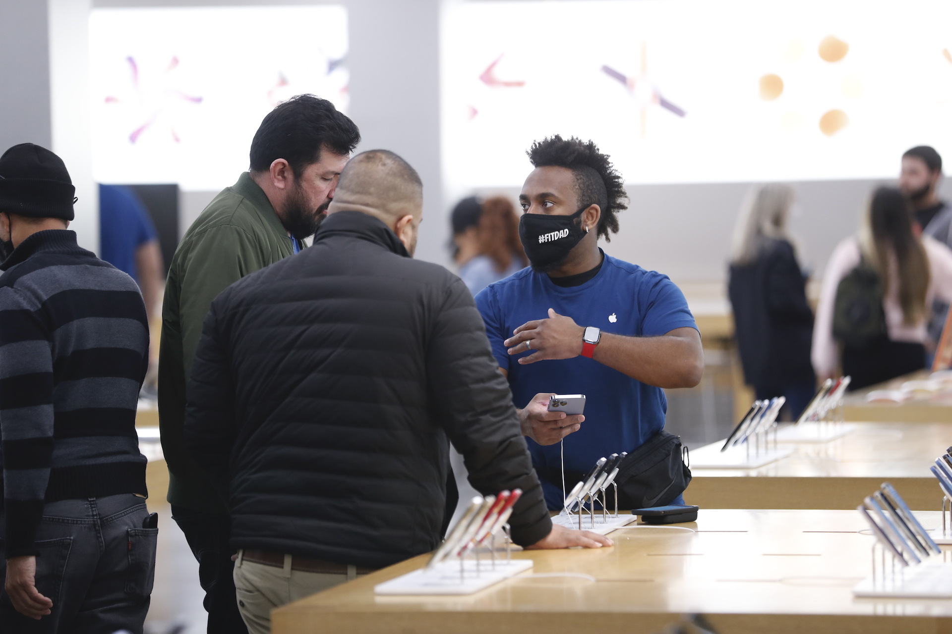 An employee in a blue shirt and black mask gestures as he talks to two male customers in winter jackets. They are at a table holding cell phones, with a white lit wall behind them.
