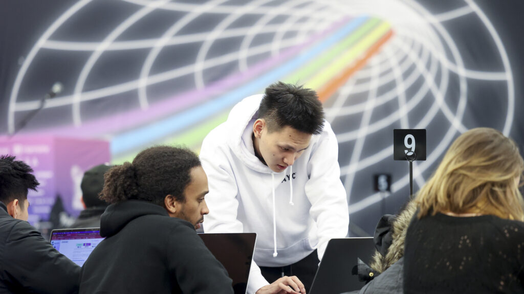 A man in a white sweatshirt bends over a laptop, while a second man sits and looks at his own computer. Behind them is a mural on the wall of a rainbow traveling through a cone.