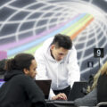 A man in a white sweatshirt bends over a laptop, while a second man sits and looks at his own computer. Behind them is a mural on the wall of a rainbow traveling through a cone.
