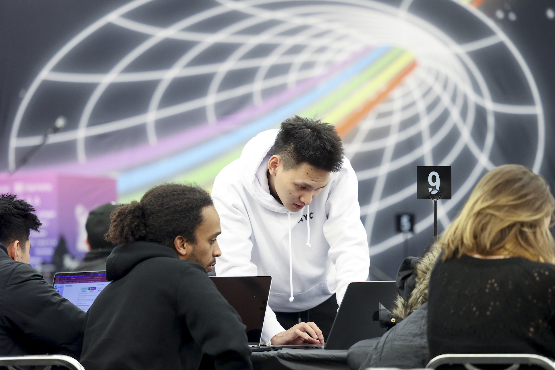 A man in a white sweatshirt bends over a laptop, while a second man sits and looks at his own computer. Behind them is a mural on the wall of a rainbow traveling through a cone.