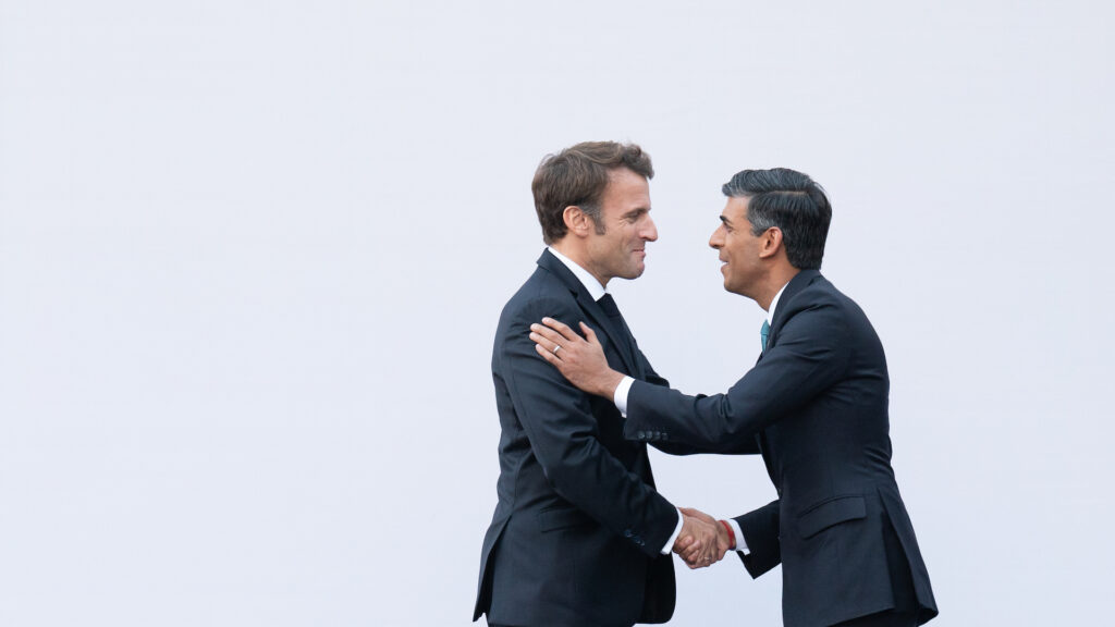 Two men in suits shake hands. Behind them is a grey wall.