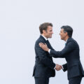 Two men in suits shake hands. Behind them is a grey wall.