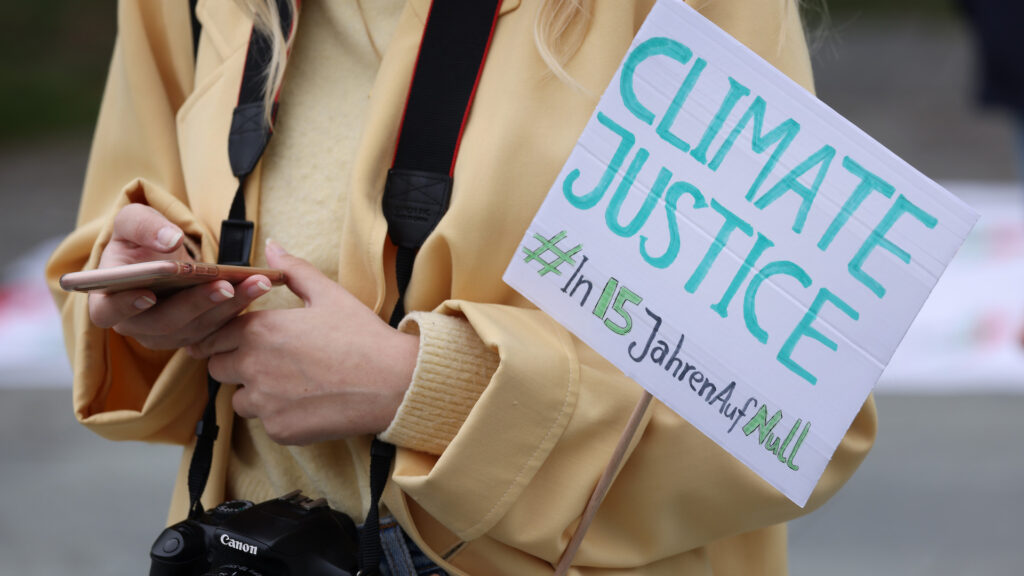 A photo of a woman's torso. She has a camera around her neck and is holding her smartphone and a sign that says in English and German, 