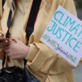 A photo of a woman's torso. She has a camera around her neck and is holding her smartphone and a sign that says in English and German, 