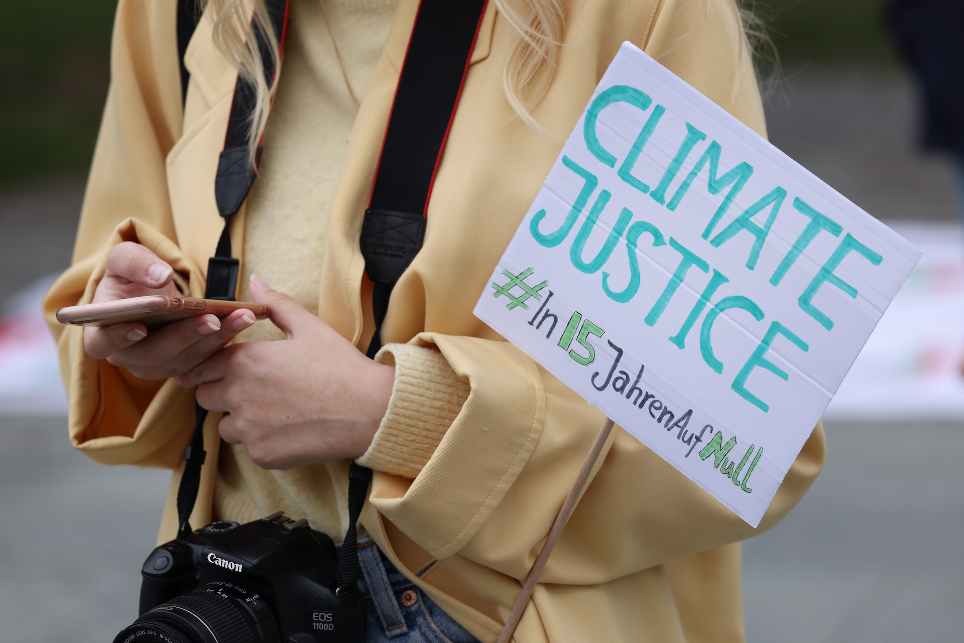 A photo of a woman's torso. She has a camera around her neck and is holding her smartphone and a sign that says in English and German,
