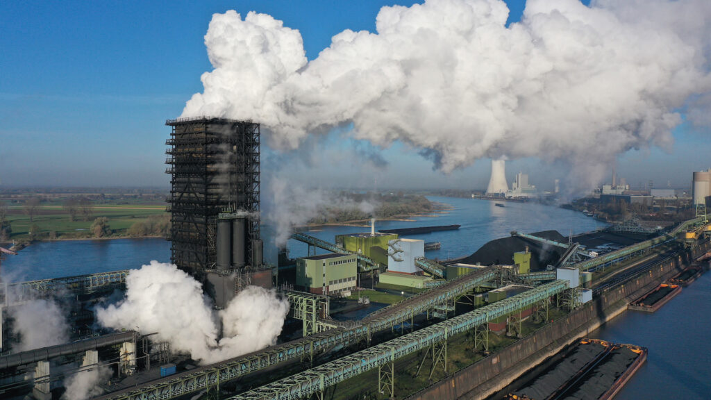 A plant near water has a large black smoke stack that belches plumes of white smoke.