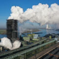 A plant near water has a large black smoke stack that belches plumes of white smoke.