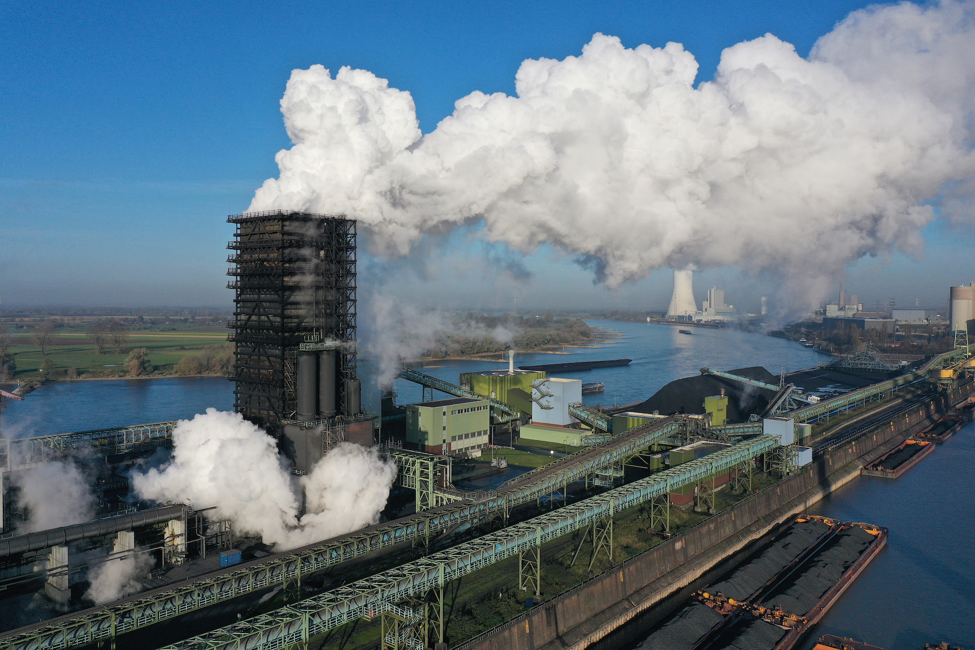 A plant near water has a large black smoke stack that belches plumes of white smoke.
