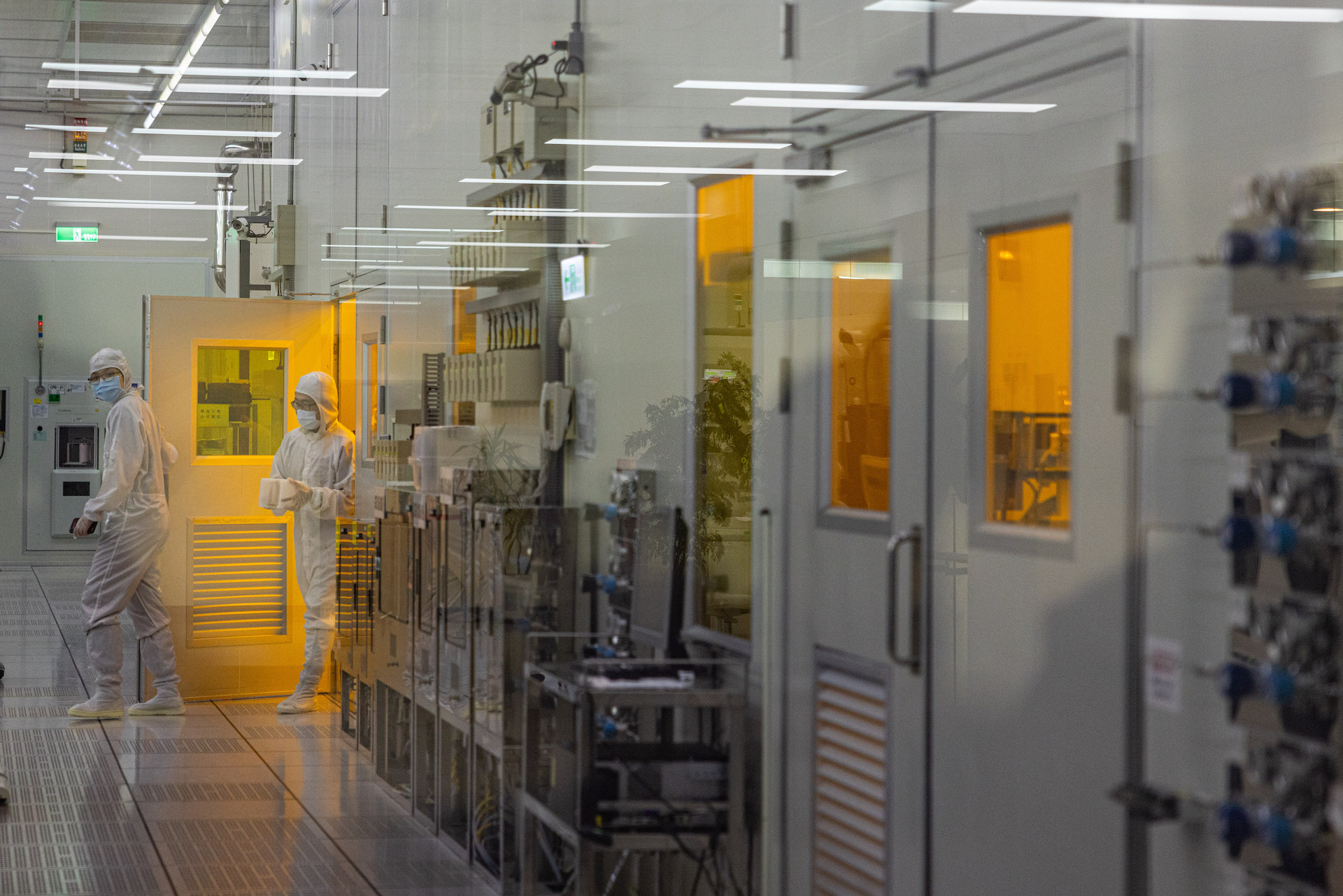 Two people wearing white suits with hoods and face masks walk through a white hallway, with yellow light spilling out of the white doors. There is glass between the camera and the subjects.