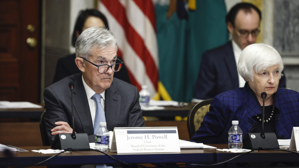 A grey-haired man in a suit, blue tie, and glasses sits with his arms crossed behind a desk, with a name plate that says Jerome H. Powell. To his left is a woman with white short hair, also in a blue suit, looking to her left . Behind them are more seated people and an American flag.