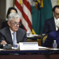 A grey-haired man in a suit, blue tie, and glasses sits with his arms crossed behind a desk, with a name plate that says Jerome H. Powell. To his left is a woman with white short hair, also in a blue suit, looking to her left . Behind them are more seated people and an American flag.