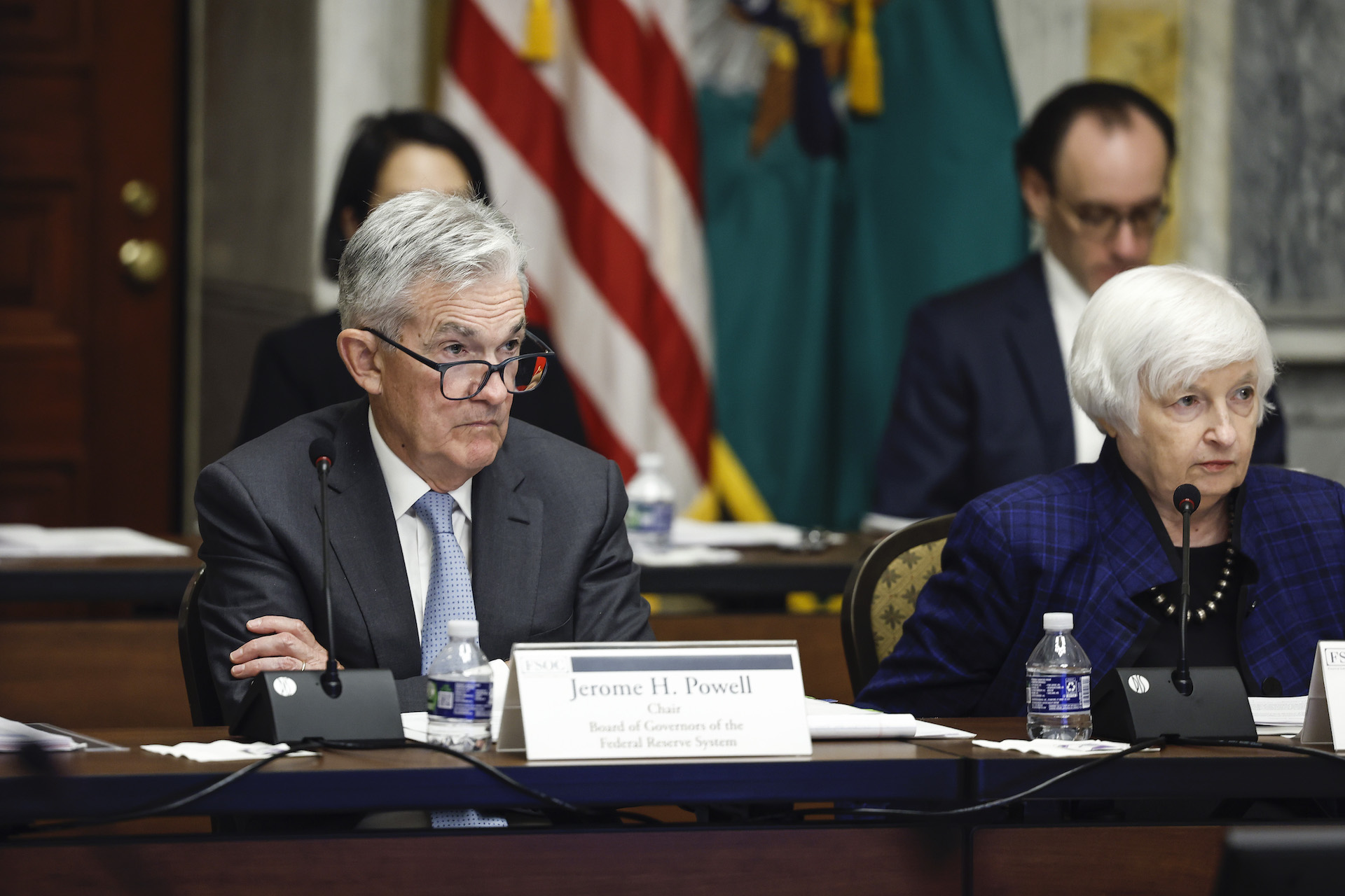 A grey-haired man in a suit, blue tie, and glasses sits with his arms crossed behind a desk, with a name plate that says Jerome H. Powell. To his left is a woman with white short hair, also in a blue suit, looking to her left . Behind them are more seated people and an American flag.