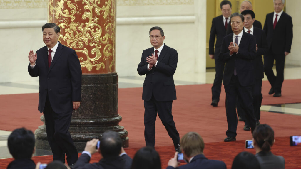 A line of men in business suits walk on a red carpeted stage as a seated audience takes pictures. There is an ornate column in the background, with a black marble base, with stylized gold flowers on a red background.