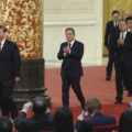 A line of men in business suits walk on a red carpeted stage as a seated audience takes pictures. There is an ornate column in the background, with a black marble base, with stylized gold flowers on a red background.