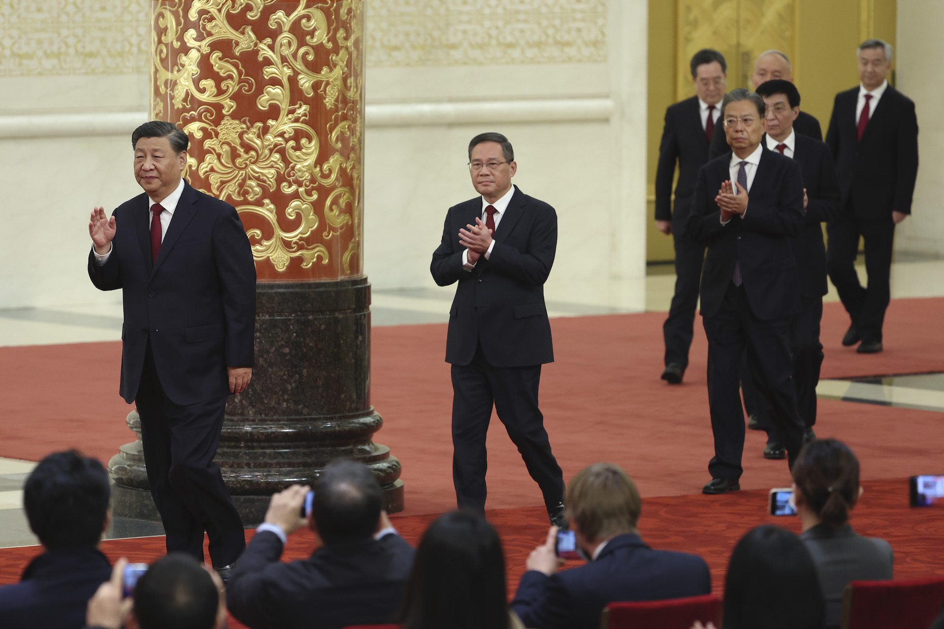 A line of men in business suits walk on a red carpeted stage as a seated audience takes pictures. There is an ornate column in the background, with a black marble base, with stylized gold flowers on a red background.