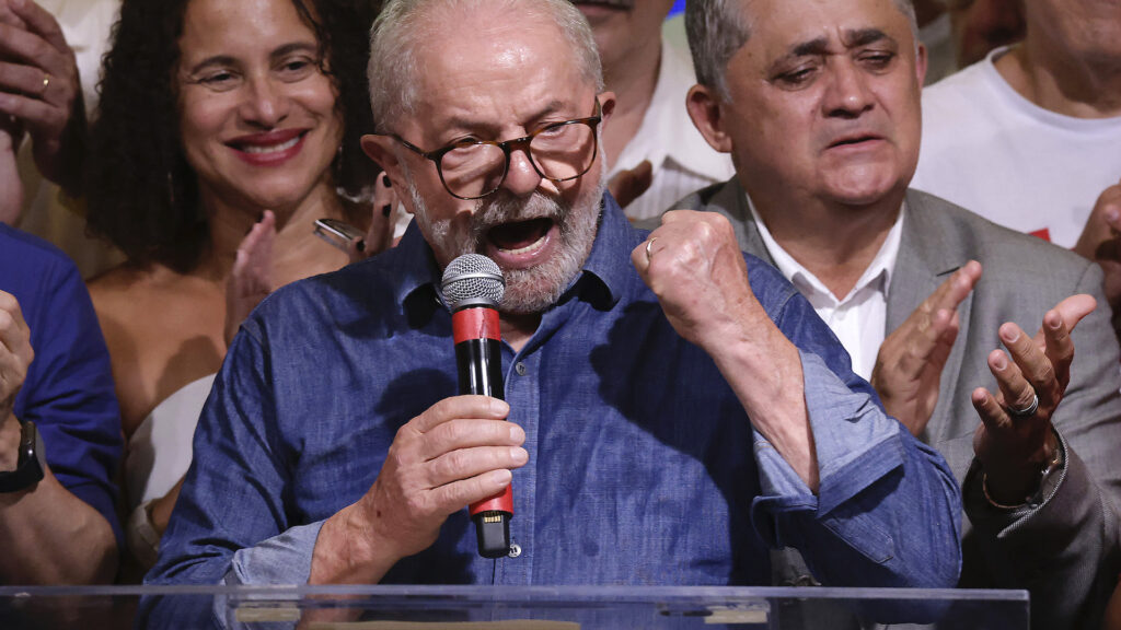 Lula holds a microphone and pumps his fist as he speaks. He stands behind a podium, and supporters are behind him. He wears a blue collared shirt and glasses.