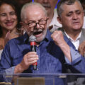 Lula holds a microphone and pumps his fist as he speaks. He stands behind a podium, and supporters are behind him. He wears a blue collared shirt and glasses.